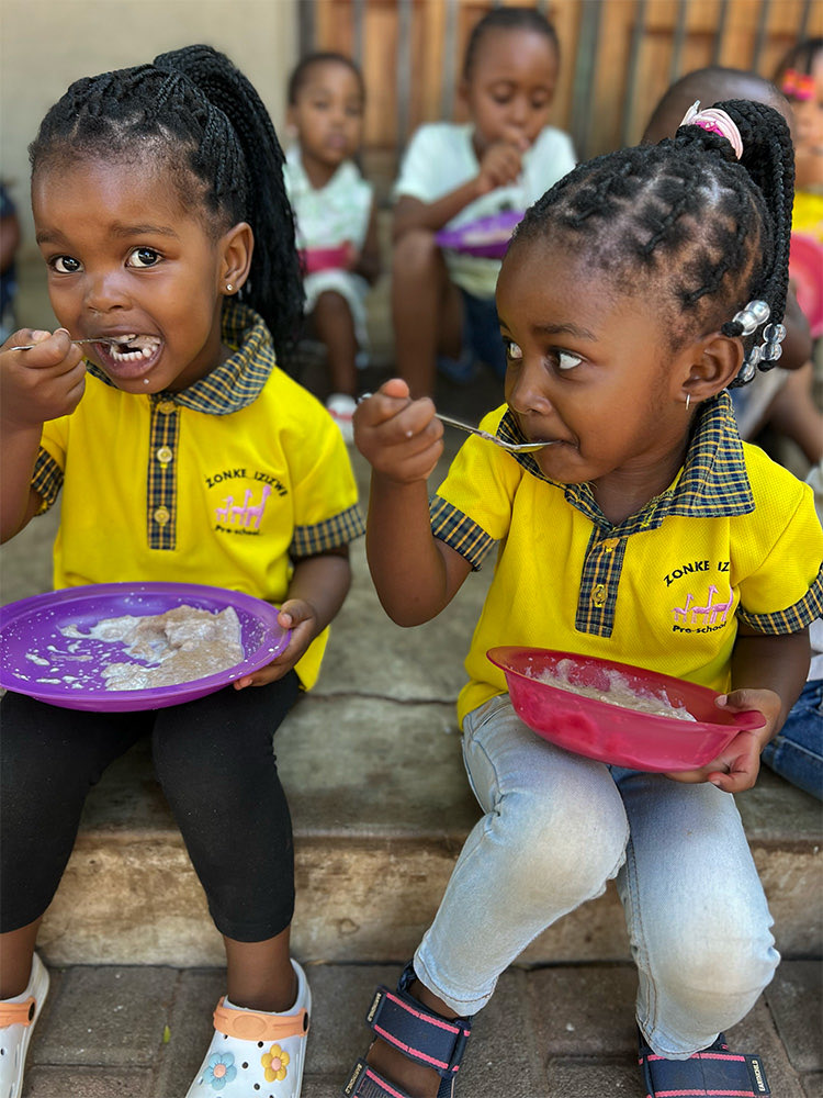 Two girls in yellow Zonke Izizwe uniforms eating porridge from coloured bowls