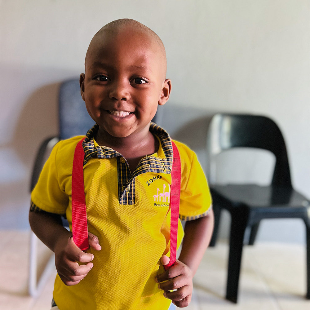 A smiling boy in a yellow Zonke Izizwe uniform wearing a red backpack
