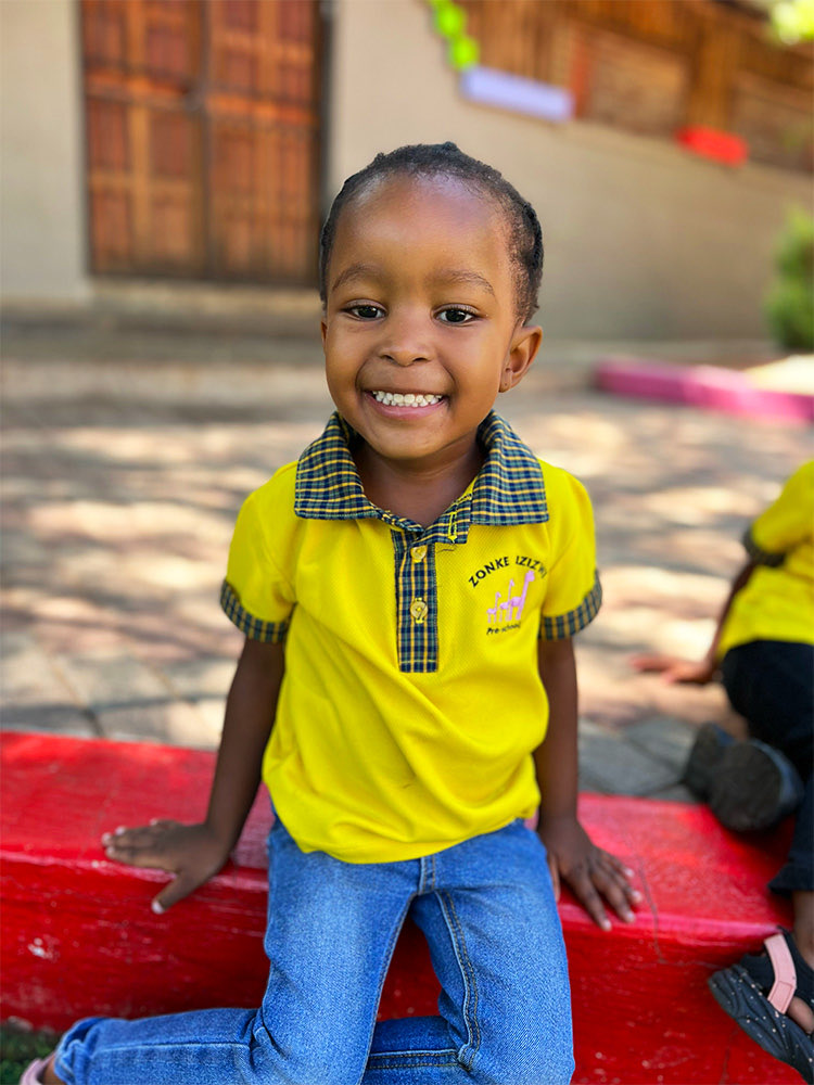 A smiling girl in a yellow Zonke Izizwe uniform sitting on a red bench